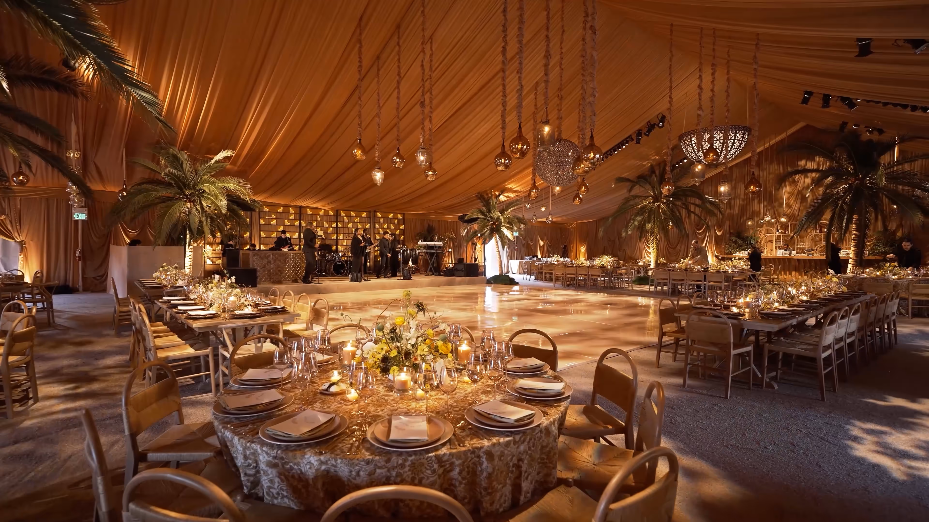 Luxurious indoor event space with round and rectangular tables set with plates, glasses, candles, floral centerpieces, palm trees, and a wooden dance floor under golden draped ceiling with hanging lights.