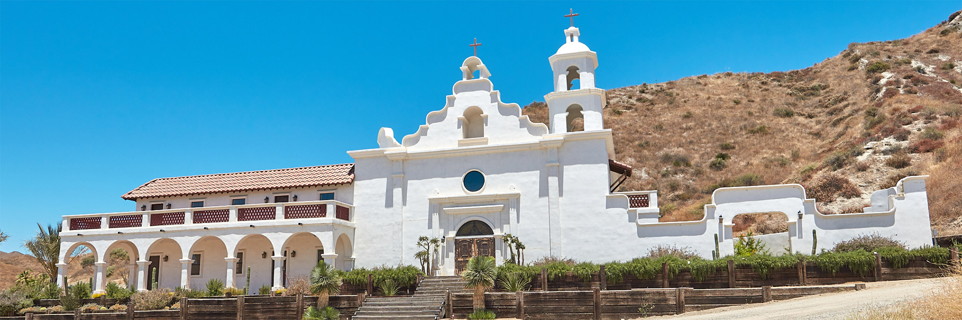 Aerial view of a small military outpost with sandbag walls on a grassy hill surrounded by rolling green mountains under a partly cloudy sky.