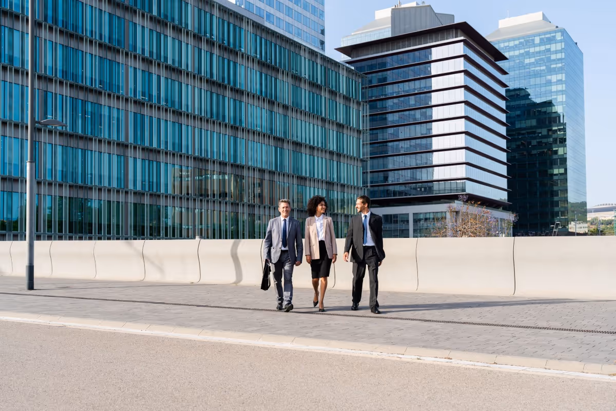 Three business professionals walking and talking on a sidewalk in front of modern glass office buildings.
