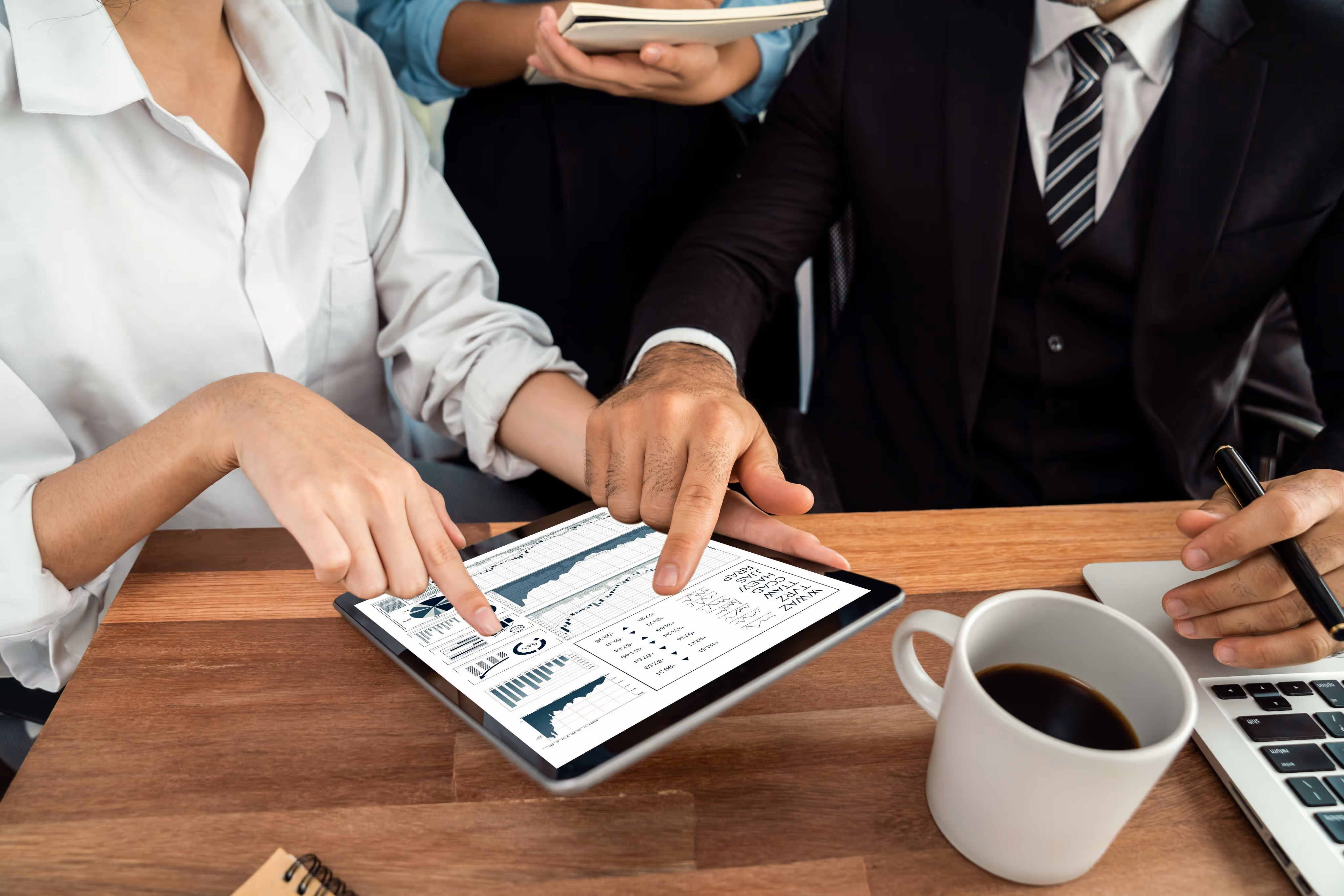 Two people pointing at financial graphs on a tablet while a third person holds a notebook nearby, with a coffee cup and laptop on the table.