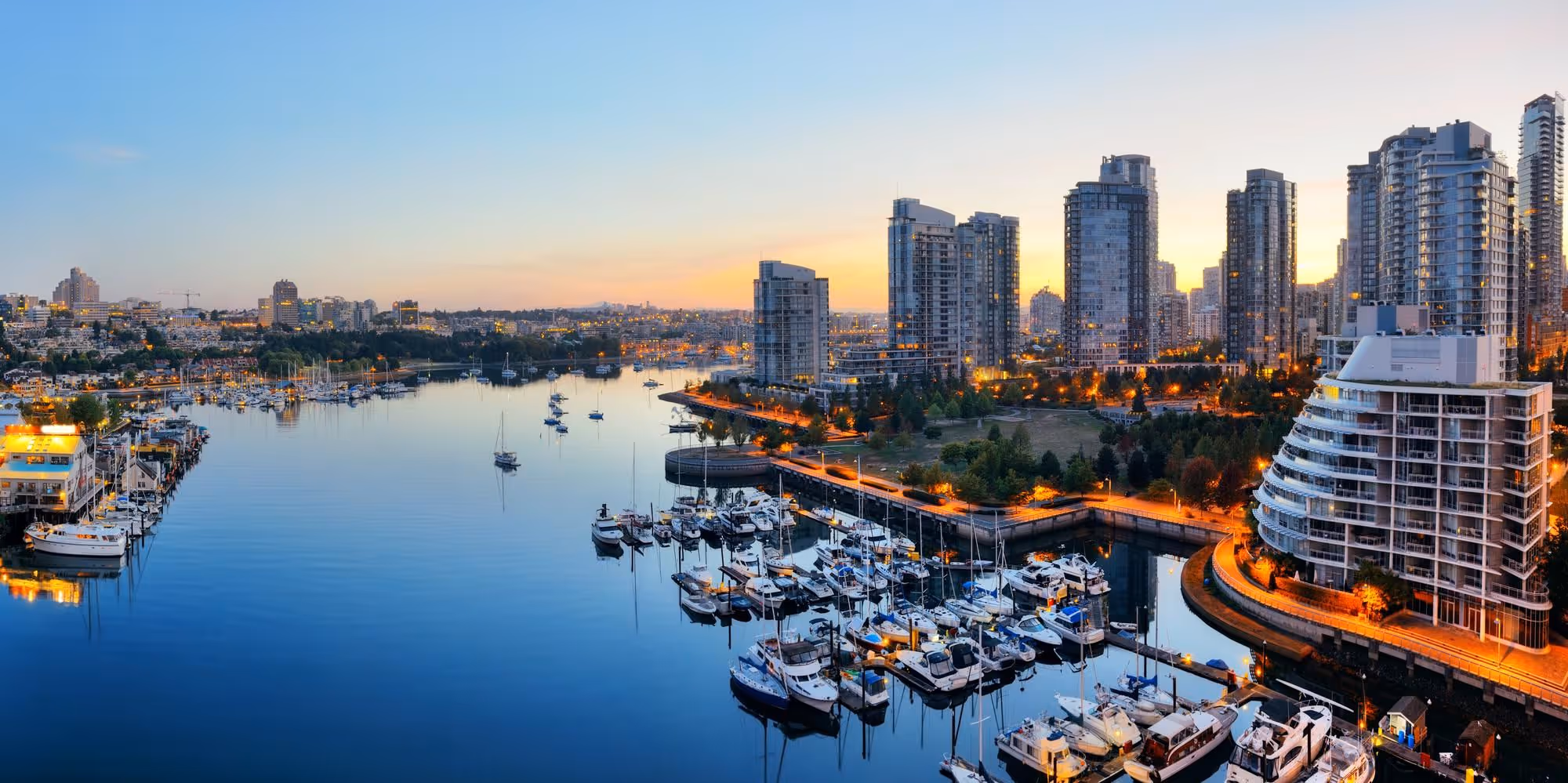 Marina filled with boats and surrounded by tall modern buildings at sunset.