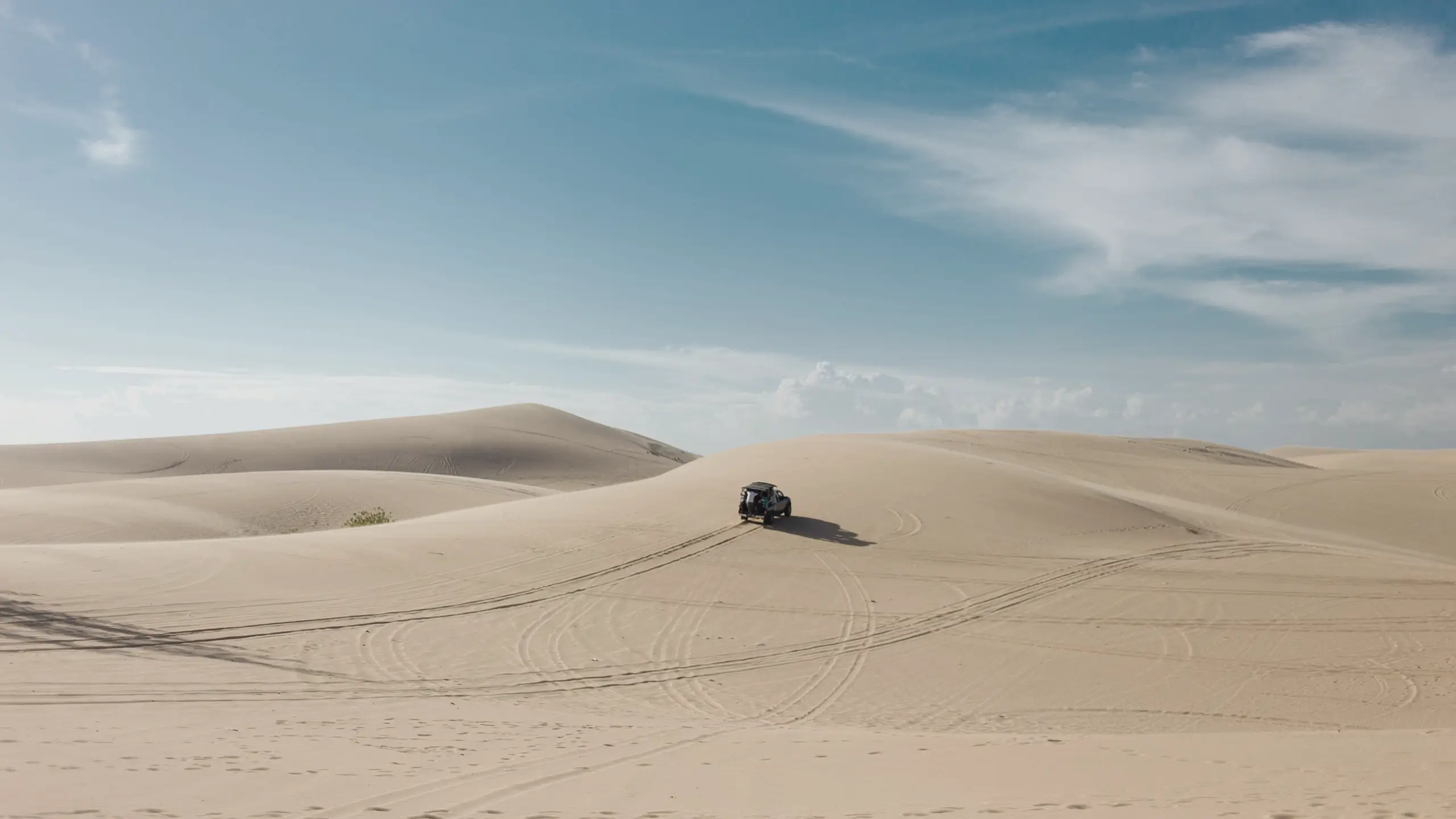 A lone off-road vehicle driving over sandy desert dunes under a blue sky with scattered clouds.