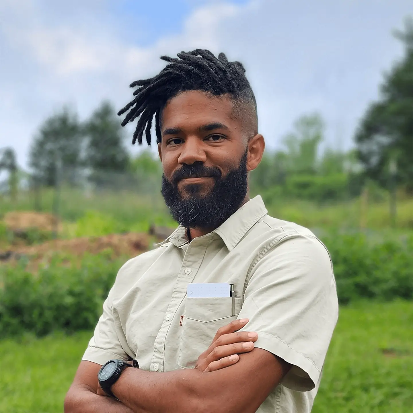 Man with dreadlocks and beard in beige shirt with arms crossed, standing outdoors with green foliage and cloudy sky in background.