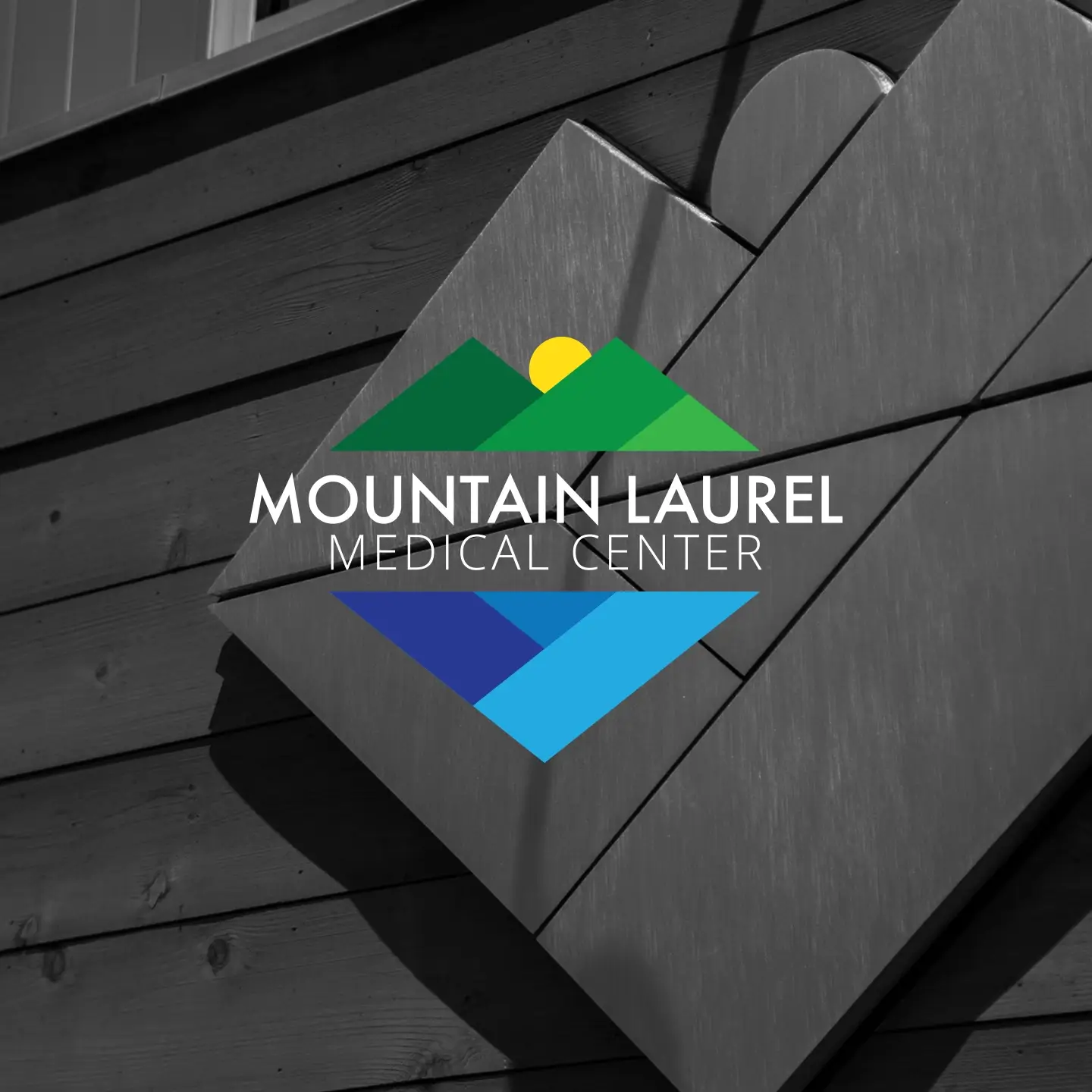 Mountain Laurel Medical Center logo featuring stylized green mountains with a yellow sun above and blue geometric shapes below on a wooden background.