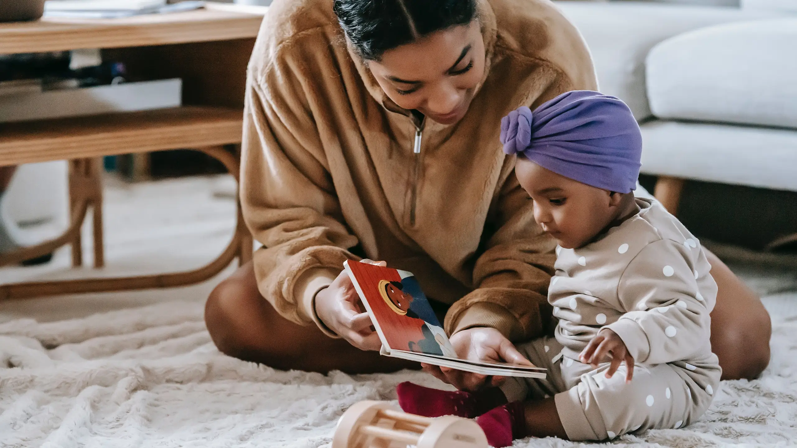 Adult reading a colorful board book to a toddler wearing a purple head wrap and polka dot pajamas on a carpeted floor.