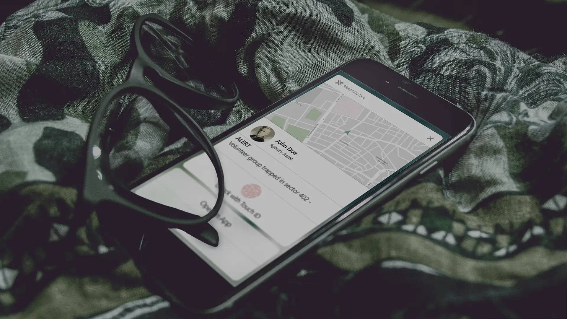 Smartphone displaying a map with an alert message about a volunteer group trapped in sector 402, next to black eyeglasses on camo fabric.