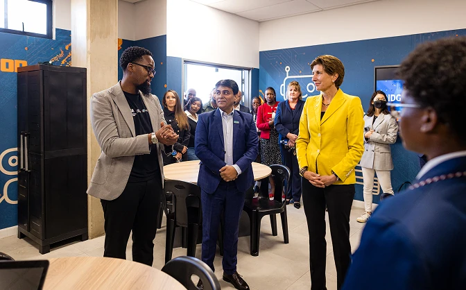 A man in a gray blazer speaking to a group including a man in a blue suit and a woman in a yellow blazer in a modern office space.