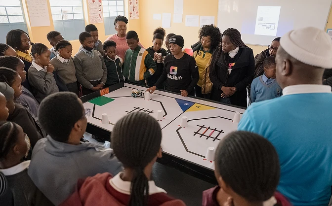 Group of students and adults gathered around a table watching a robotics demonstration in a classroom.