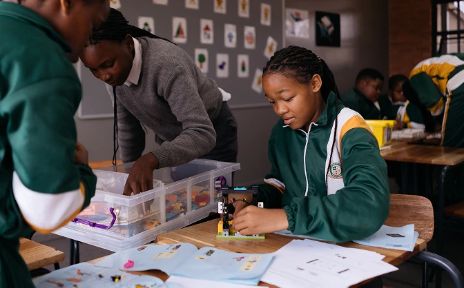 Students in green and yellow school uniforms working on a hands-on building project with instruction sheets in a classroom.