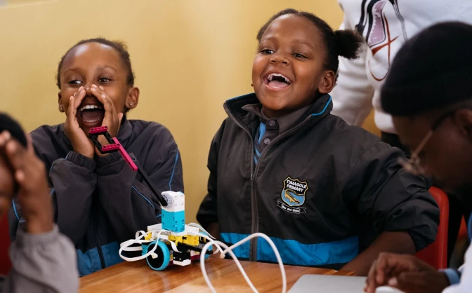 Two children laughing and engaged with a small robot on a table during a classroom activity.