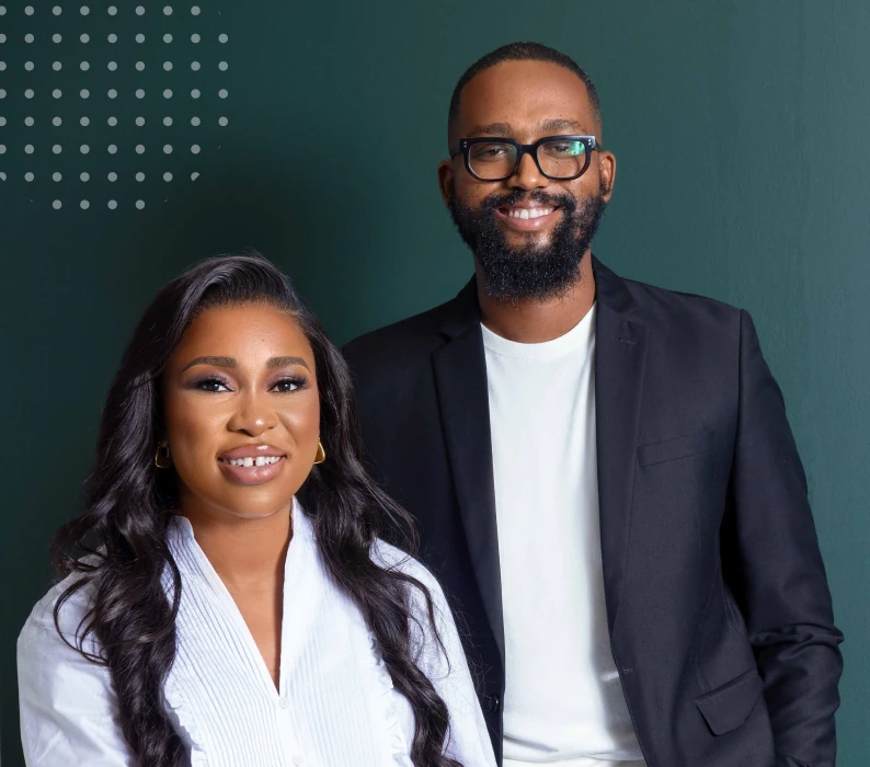 Portrait of a smiling man with glasses and beard wearing a black blazer and white shirt, and a smiling woman with long wavy hair in a white blouse standing against a dark background.