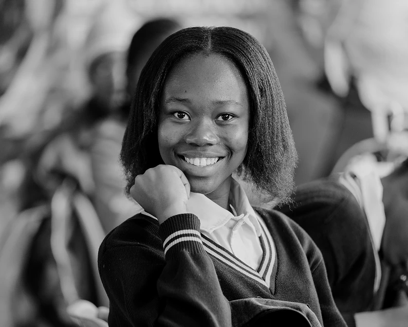 Smiling girl in a school uniform sitting with her hand resting on her chin in a classroom.
