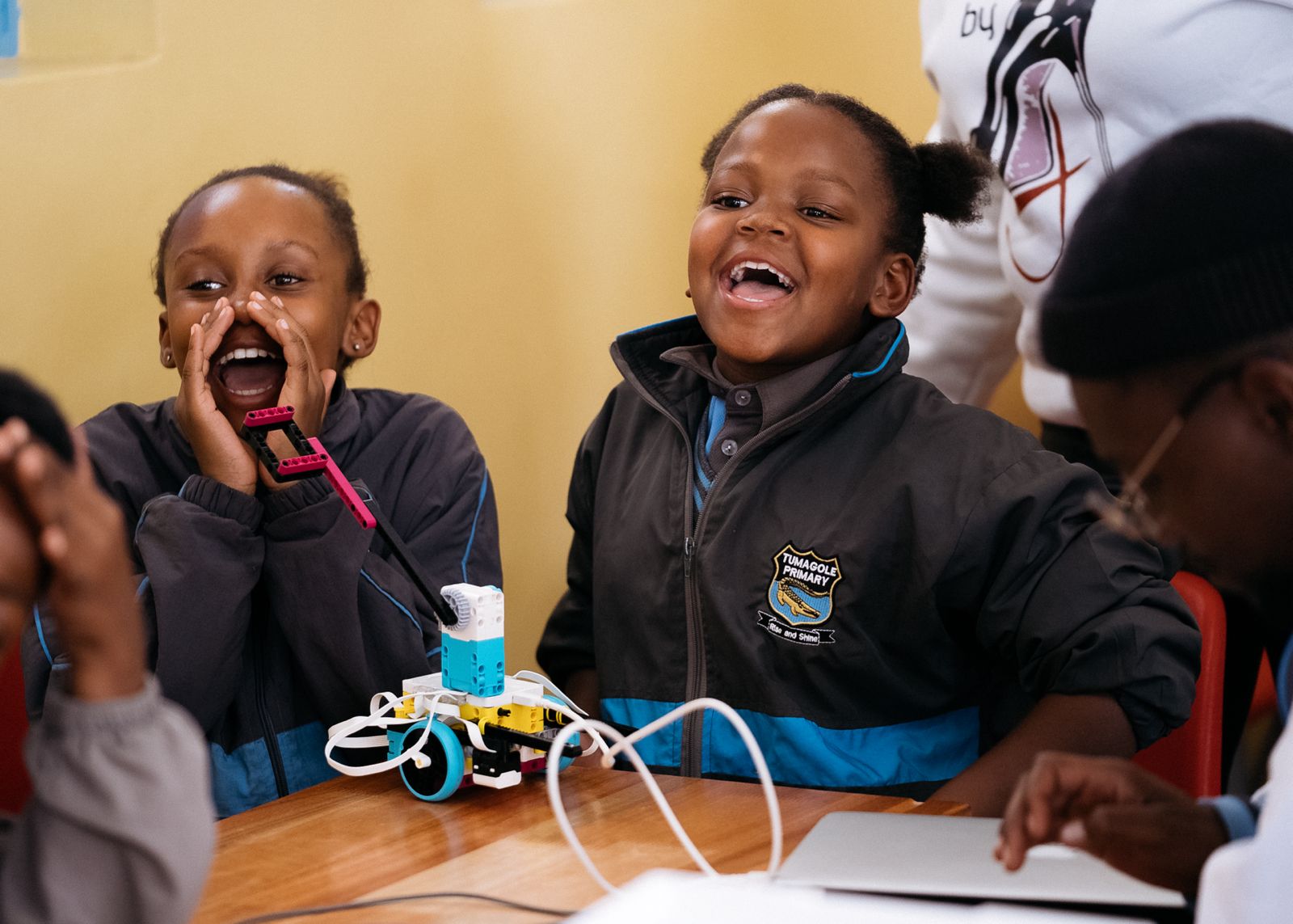 Two girls laughing and enjoying a robotic project at a table, with one boy focused on a laptop nearby.