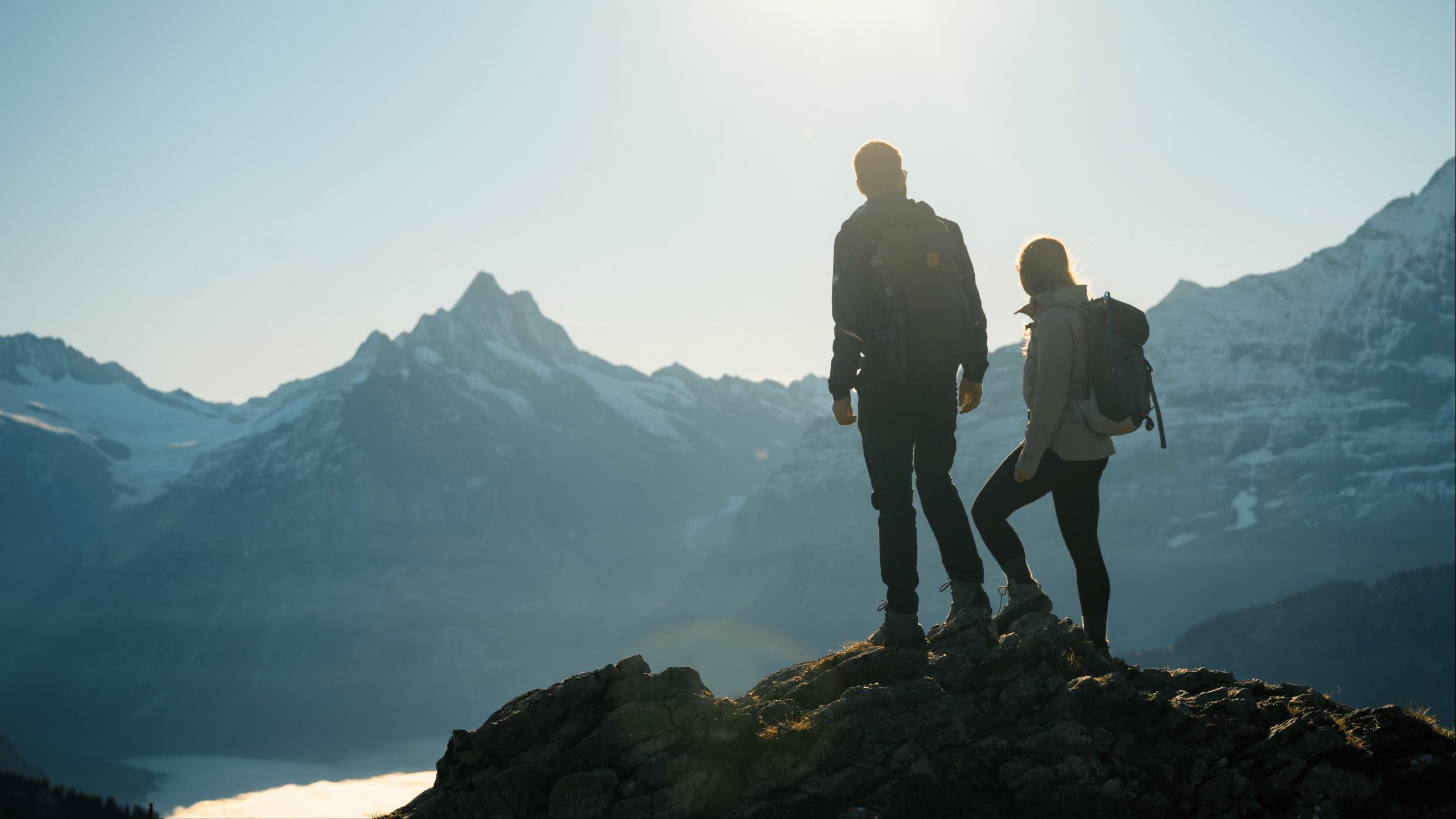 Zwei Wanderer mit Rucksäcken stehen auf einem Felsen und blicken auf schneebedeckte Berge bei Sonnenaufgang.