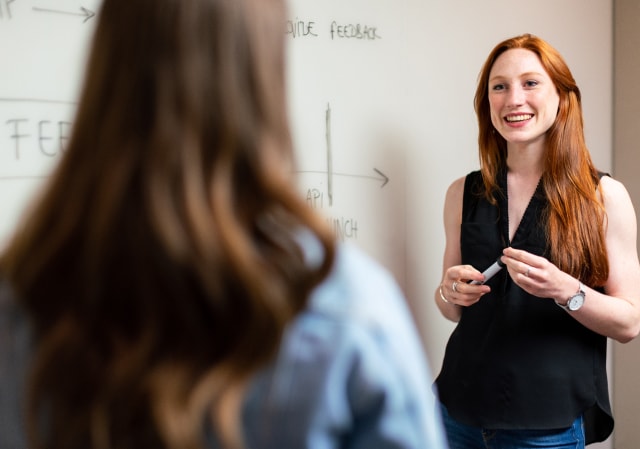 Vrouw met rood haar die glimlachend een marker vasthoudt tijdens het uitleggen voor een whiteboard.