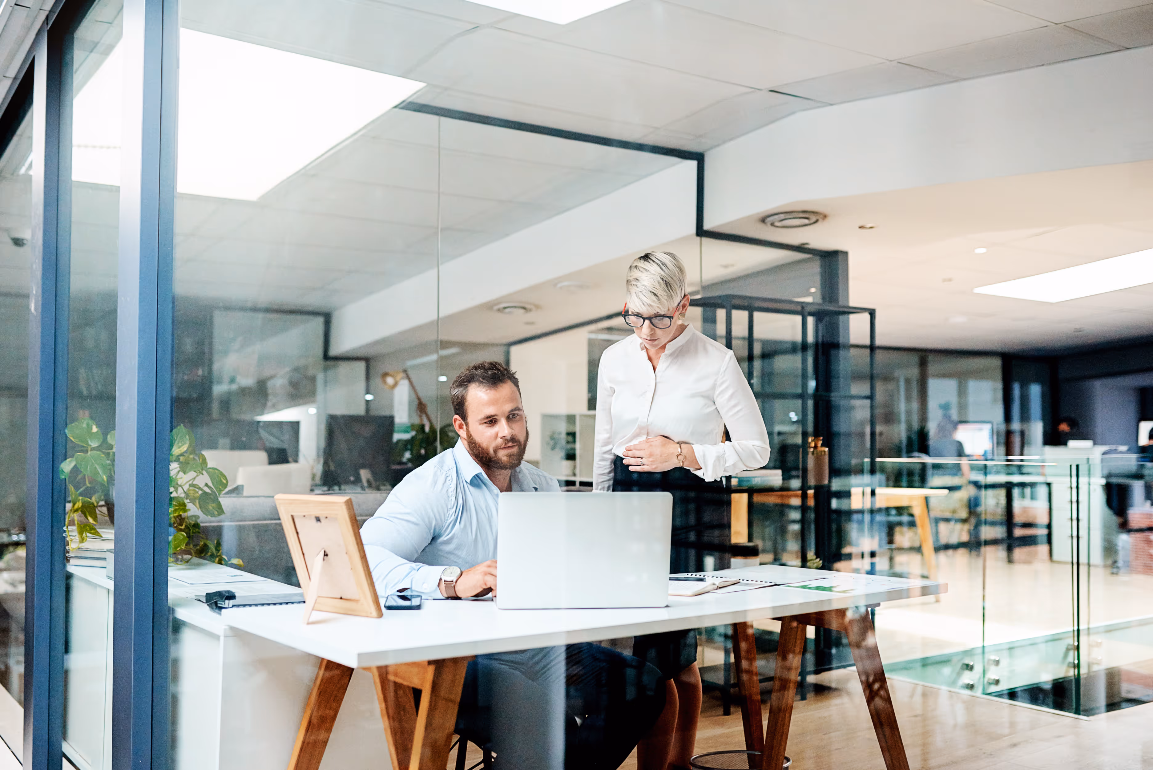 Twee collega's werken samen aan een laptop in een modern kantoor met glazen wanden.