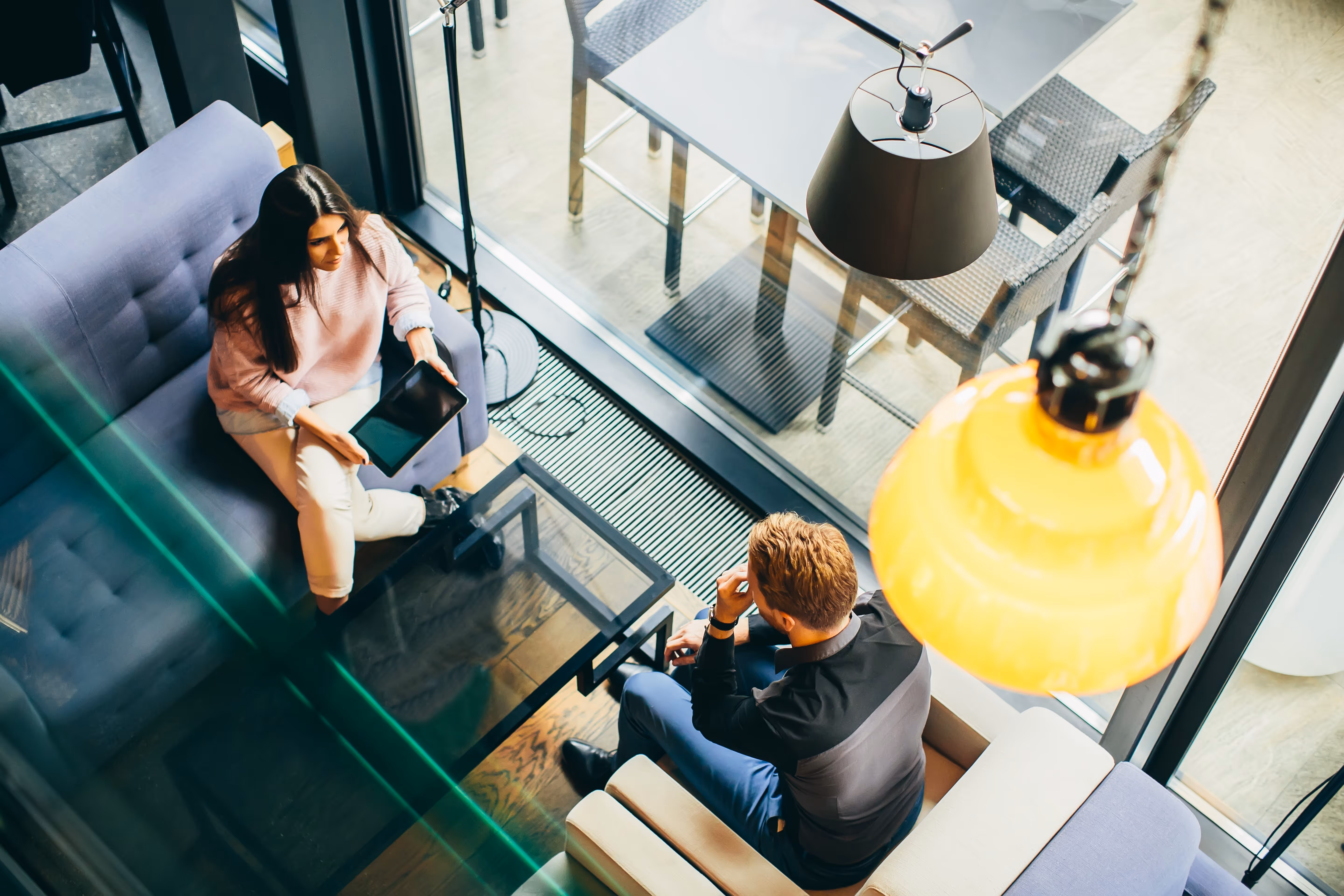 Twee jonge volwassenen zitten in een moderne zithoek, met een vrouw die een tablet vasthoudt en een man die tegenover haar zit.