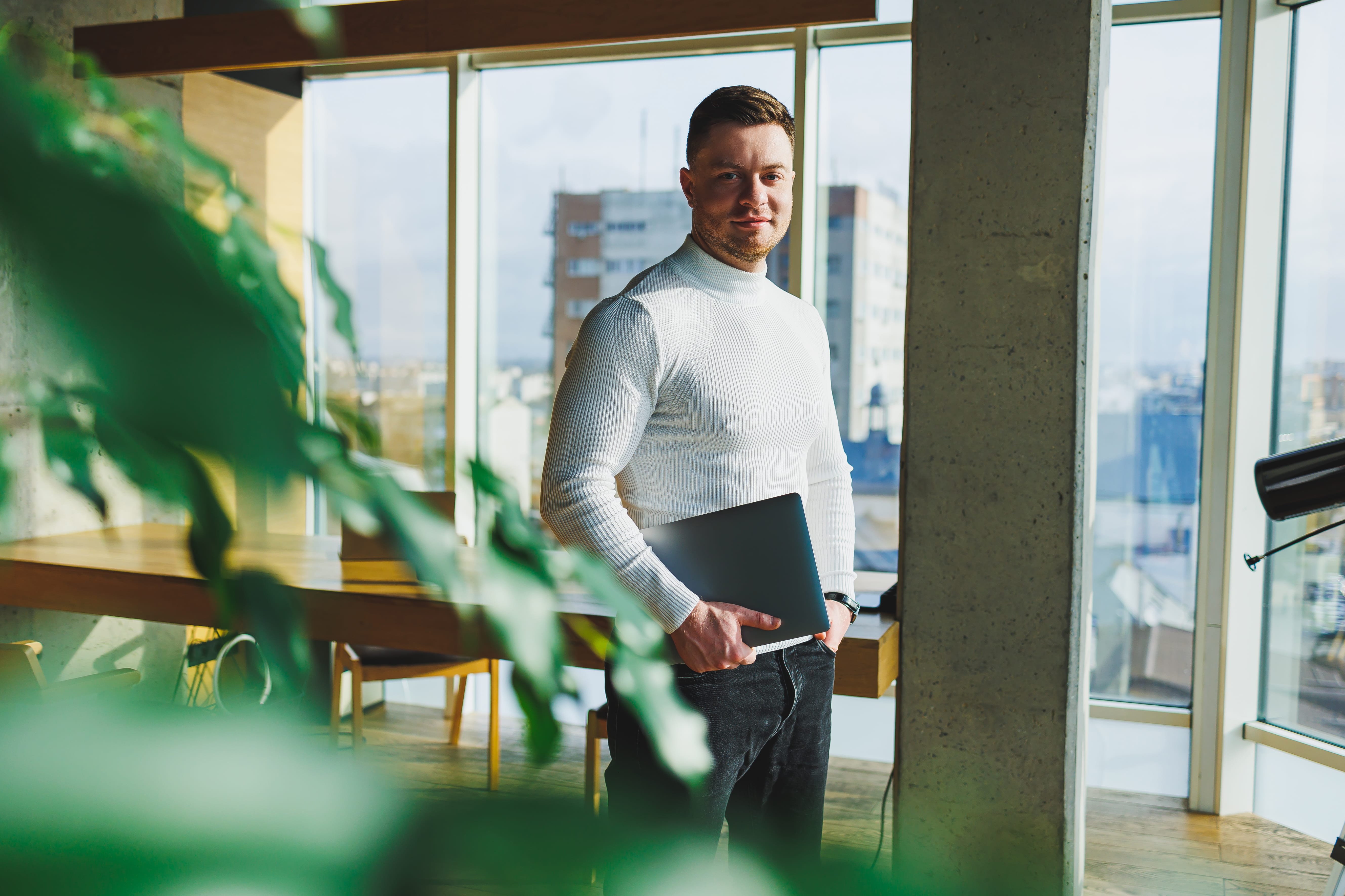Man met witte coltrui met laptop onder zijn arm in een modern kantoor met grote ramen en stadsuitzicht.