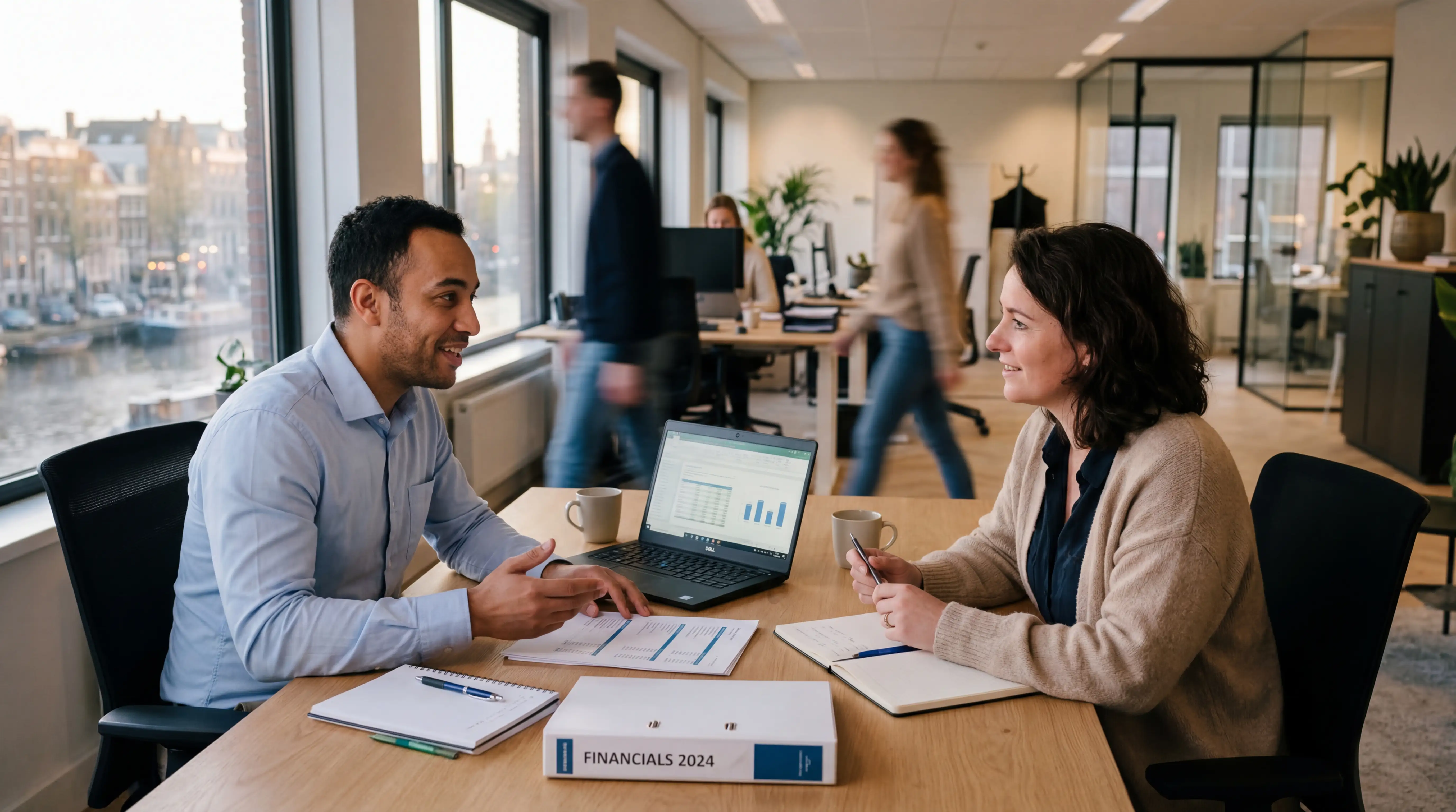 Twee collega's overleggen aan een bureau met een open laptop en documenten in een modern kantoor met uitzicht op een kanaal.