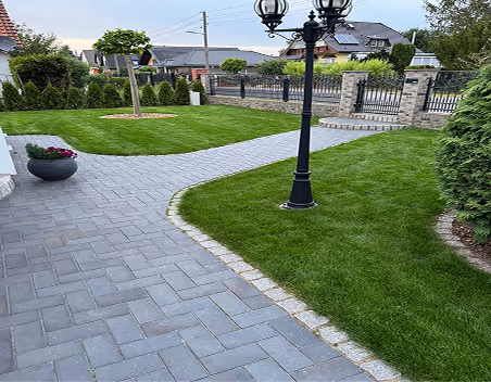 Gepflasterter Weg führt durch einen gepflegten Garten mit grünem Rasen, Baum, Laternenpfahl und Häusern im Hintergrund.