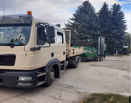 Beiges Lastwagen-Cab zieht einen Anhänger mit grünem Forstschredder auf Asphaltfläche vor Nadelbäumen.