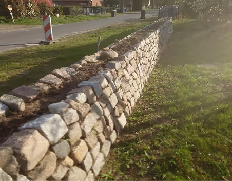 Eine lange, trockene Mauer aus unregelmäßigen Steinen neben einem grasbewachsenen Weg unter Sonnenschein.