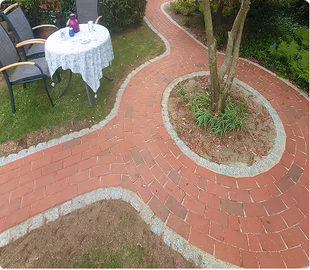 Geschwungener roter Backsteinpfad um einen Baum mit grünen Pflanzen, daneben ein mit weißer Tischdecke gedeckter Gartentisch und Stühle auf Rasen.