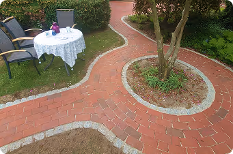 Geschwungener roter Ziegelsteinweg um einen Baum auf einem Garten mit verziertem runden Tisch und Stühlen.