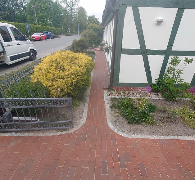 Schmaler roter Ziegelweg neben einem Fachwerkhaus mit grünen Holzbalken und Garten mit verschiedenen Pflanzen.