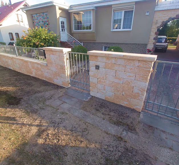 Eingangstor aus Metall und Steinmauer vor einem beige gestrichenen Einfamilienhaus mit Vorgarten.
