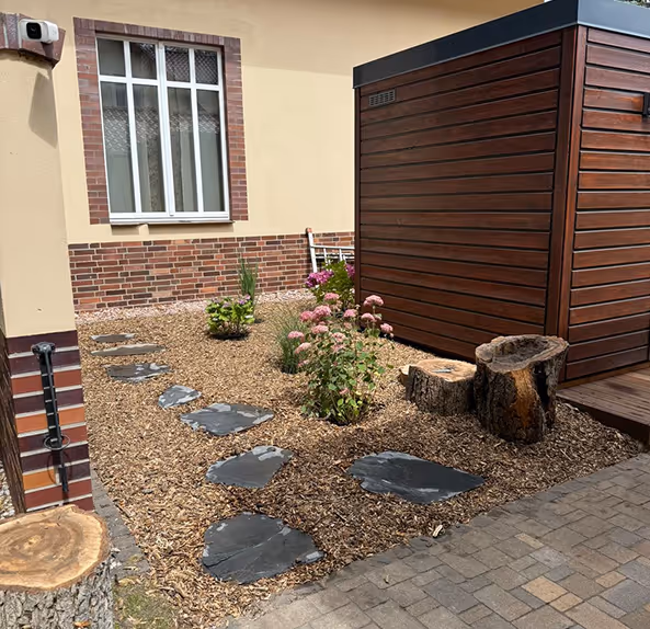 Kleiner Gartenweg mit schwarzen Steinplatten, umgeben von Holzspänen, Blumen und einem Holzhaus neben einem Haus mit gelber Fassade und weißem Fenster.