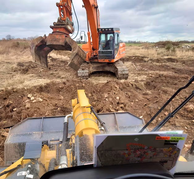 Blick aus dem Fahrerhaus eines gelben Bauwagens auf einen orangen Bagger mit Greifklaue auf einem unebenen, erdigen Baustellengelände unter bewölktem Himmel.