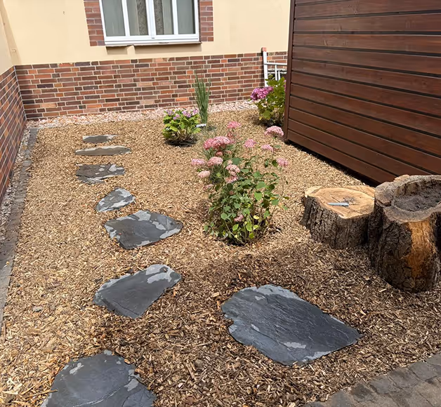 Ein gepflegter Garten mit brauner Mulchbedeckung, dunkelgrauen Trittsteinen, blühenden Pflanzen und einem Holzstamm als Dekoration neben einer braunen Holzwand.