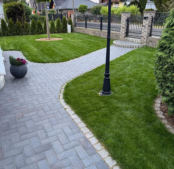Ein gepflegter Garten mit grünem Rasen, einem gepflasterten Gehweg, einer schwarzen Laterne und einer schwarzen Blumentopfvase mit roten Blumen.