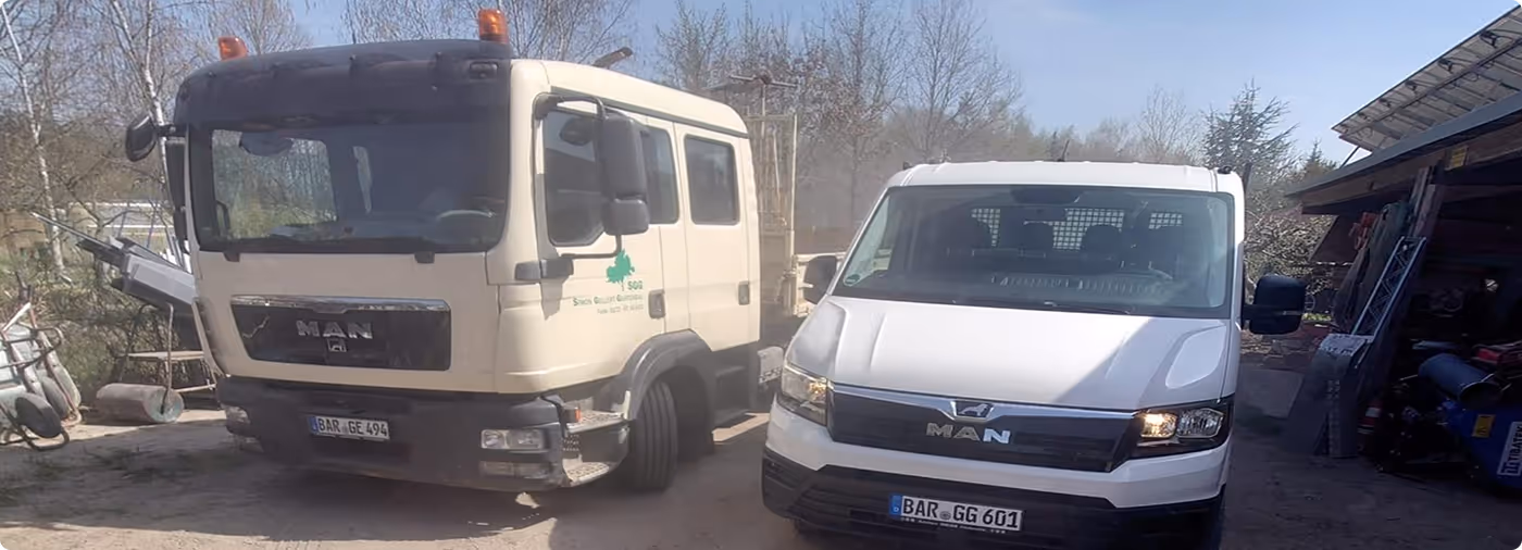 Ein weißer Lastwagen und ein weißer Lieferwagen mit MAN-Logo, geparkt nebeneinander vor einem Schuppen und Bäumen im Hintergrund.