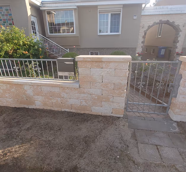 Haus mit hellen Steinmauern, einem Metallzaun mit Briefkästen und einem Tor, das zu einem gepflasterten Weg führt.