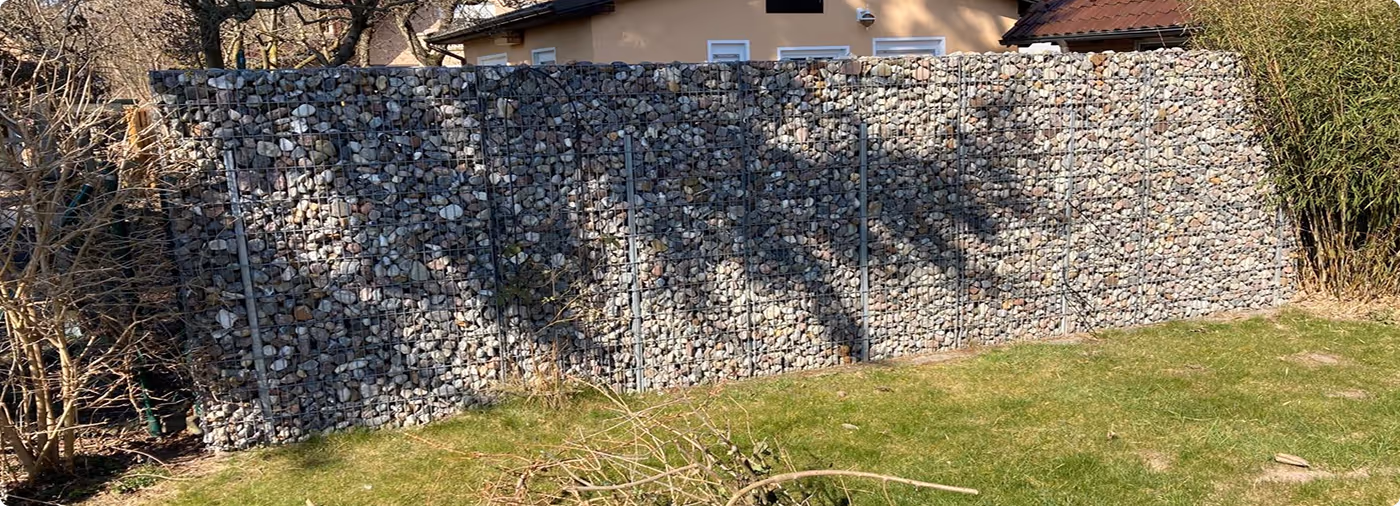 Steinmauer als Gartenbegrenzung vor einem Haus mit grünem Rasen und Schatten von Bäumen.