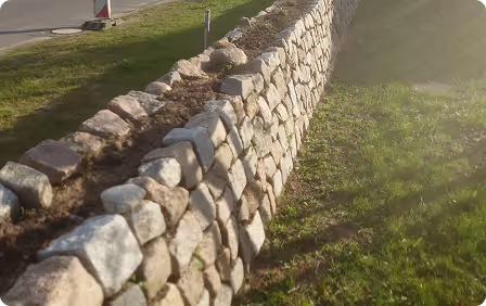 Sunlit stone retaining wall with grass on one side and a paved area on the other.