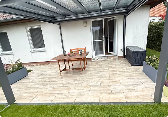 Überdachte Terrasse mit Holztisch, Bank, Pflanzenkästen und offenem Glastür zur weißen Hauswand.