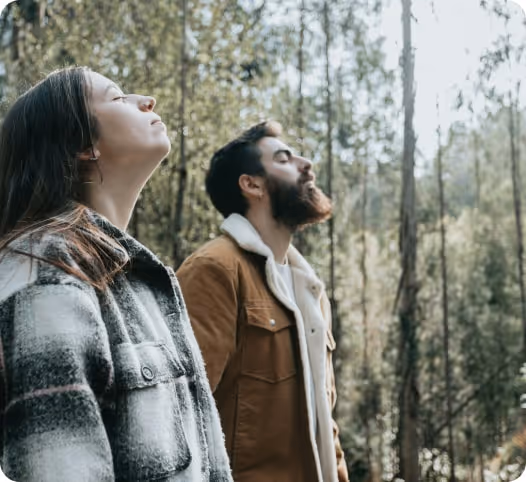 A man and a woman with eyes closed, standing outdoors in a forest, enjoying the fresh air.