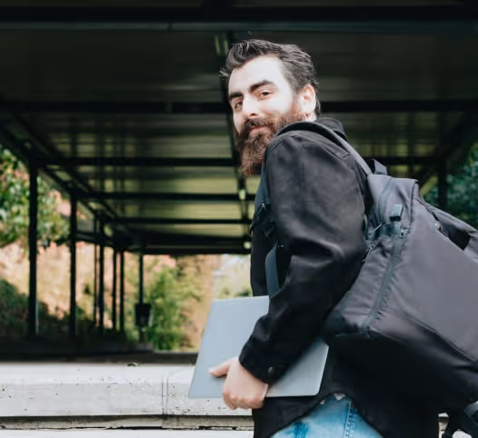 Man with a beard carrying a backpack and holding a laptop walking under a covered walkway.