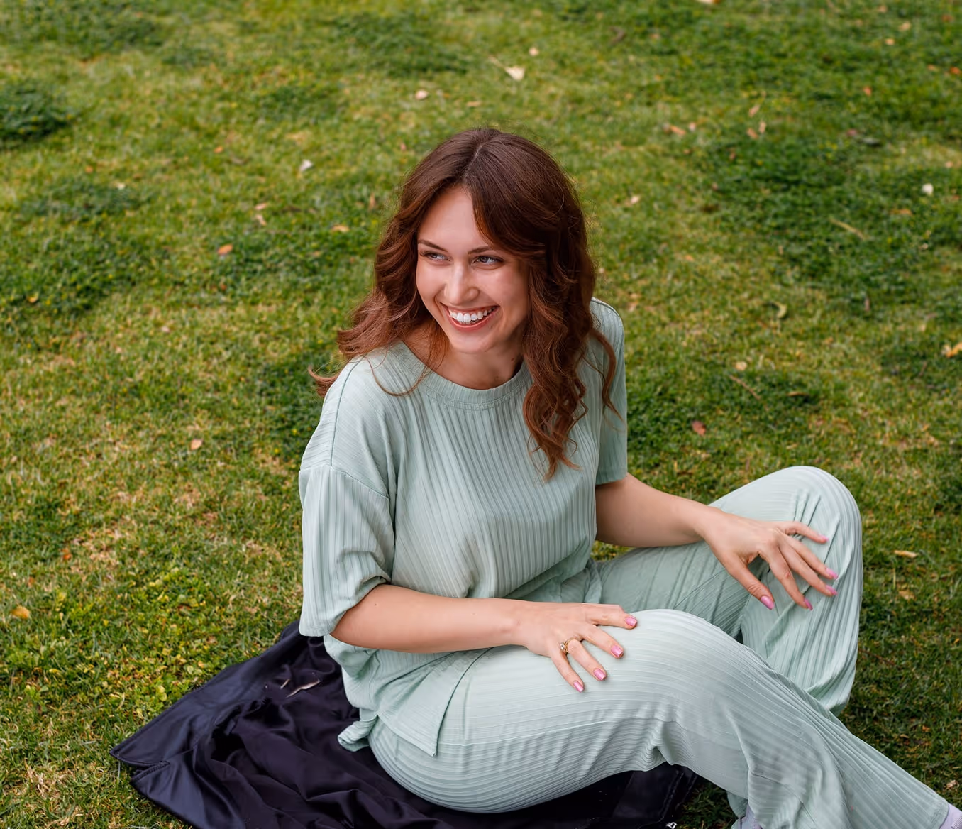 Smiling woman with wavy brown hair wearing a light green ribbed outfit sitting on grass with a dark cloth underneath.