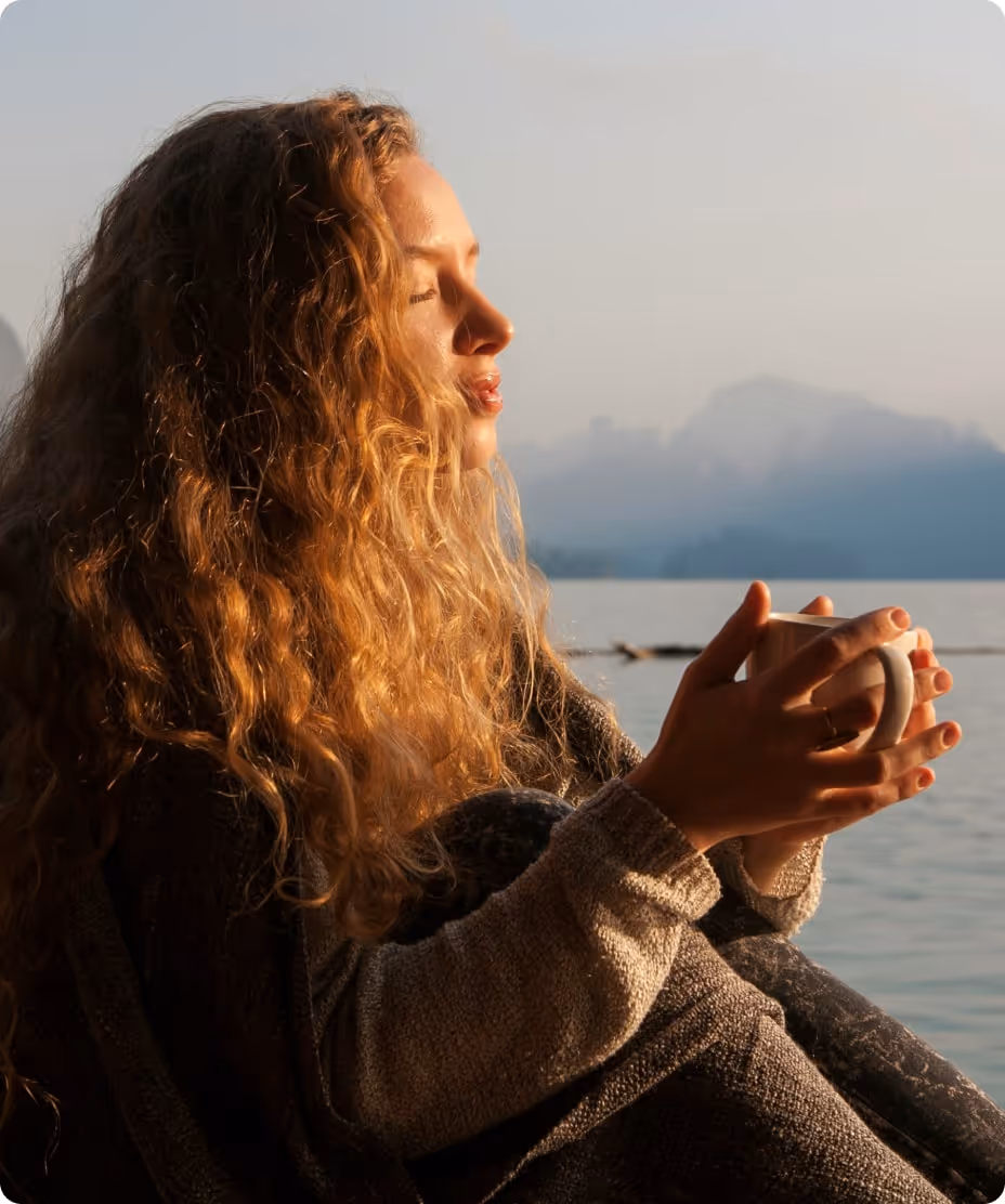 Woman with curly hair sitting by a lake at sunrise or sunset, holding a cup and enjoying the peaceful view.