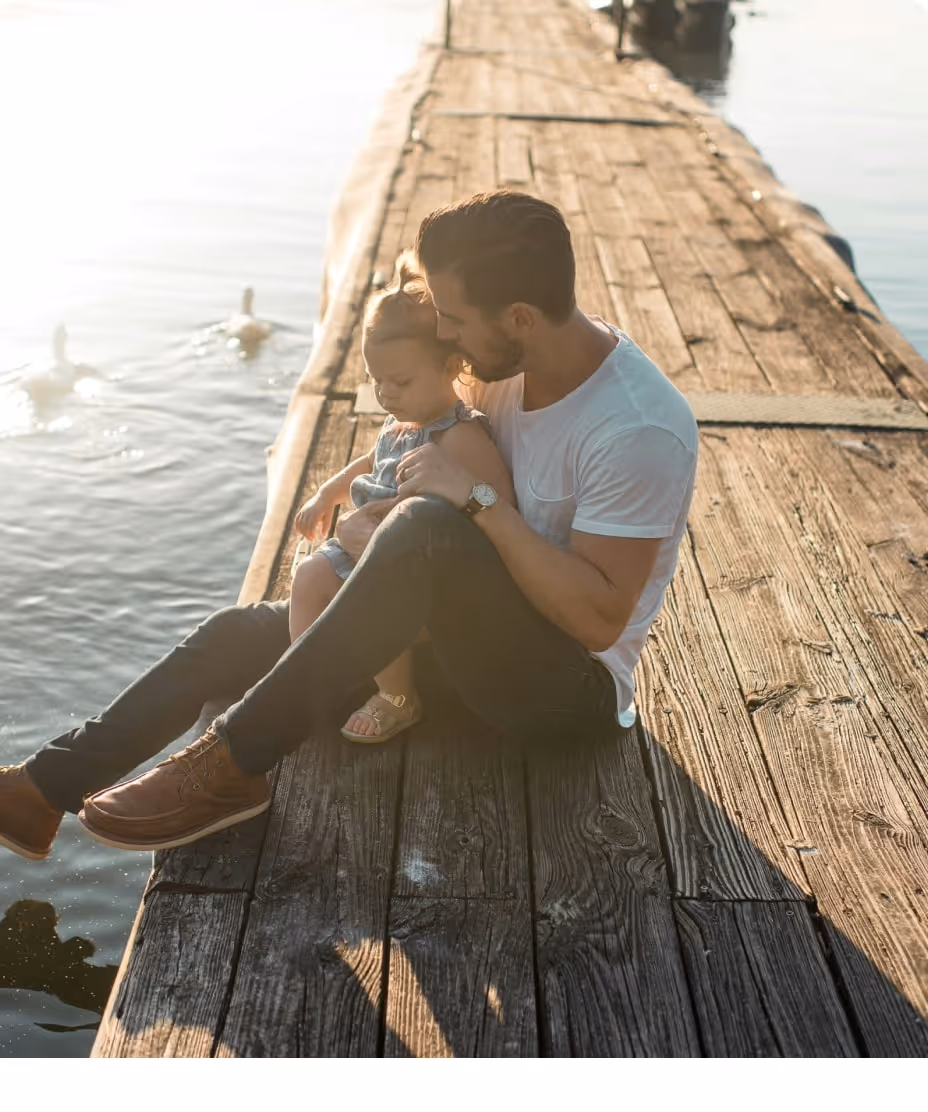 Man sitting on a wooden dock holding a young child, with water and ducks nearby in soft sunlight.