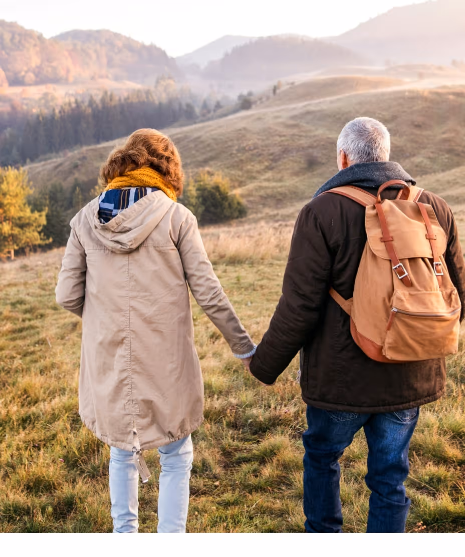 Couple holding hands and walking outdoors on grass with hills and trees in the background during autumn.