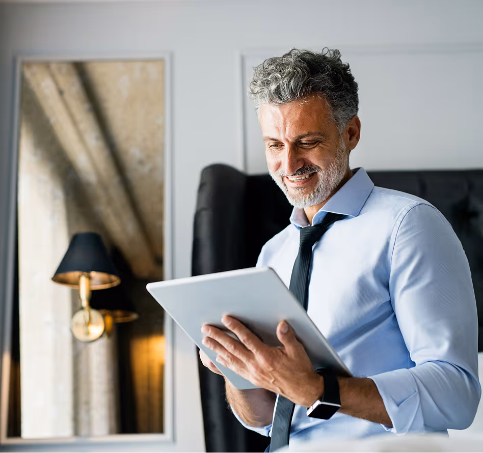 Smiling mature man with gray hair and beard using a tablet indoors, wearing a light blue shirt and black tie.