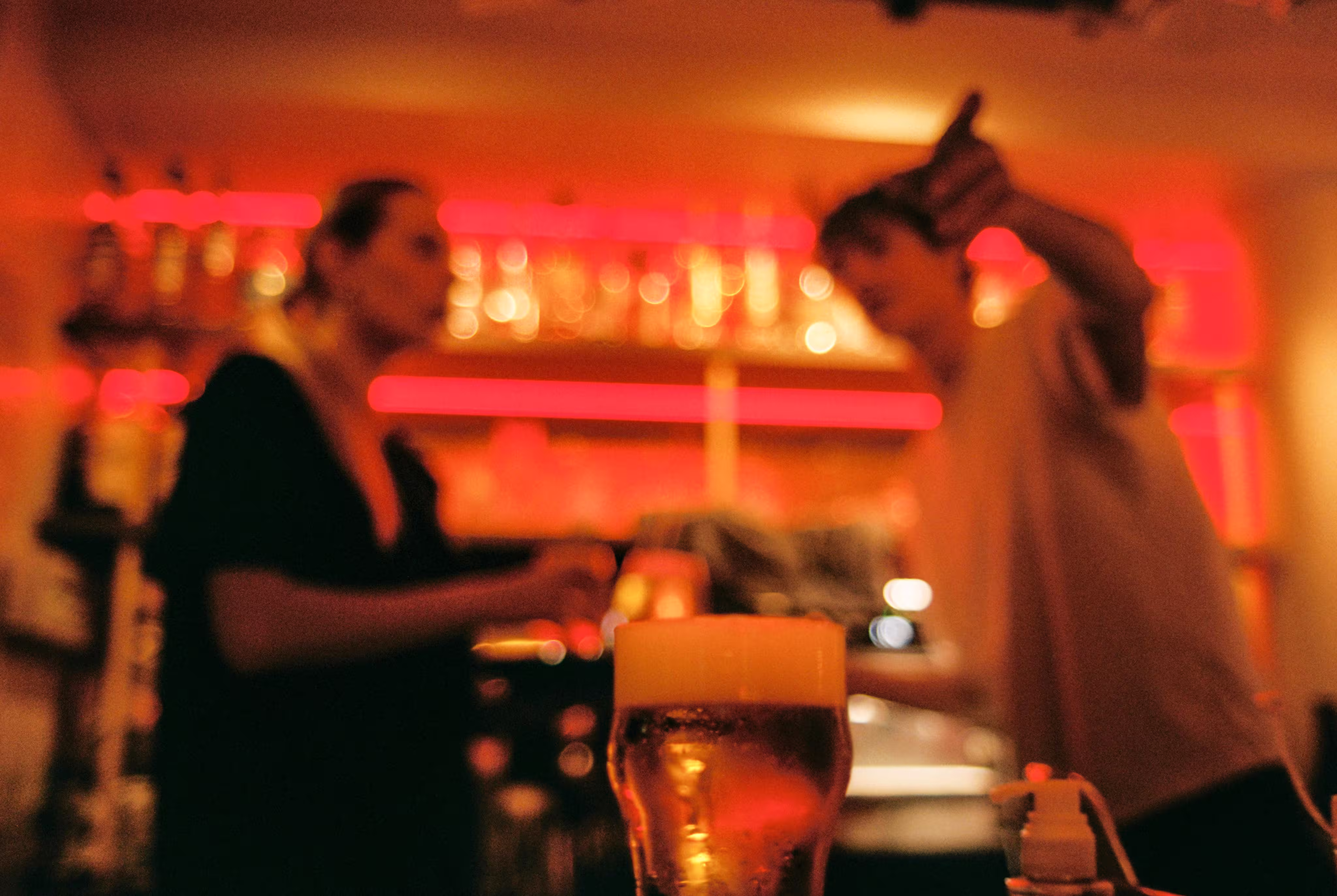 Un verre de bière mousseuse au premier plan avec deux personnes floues en arrière-plan dans un bar éclairé en rouge.