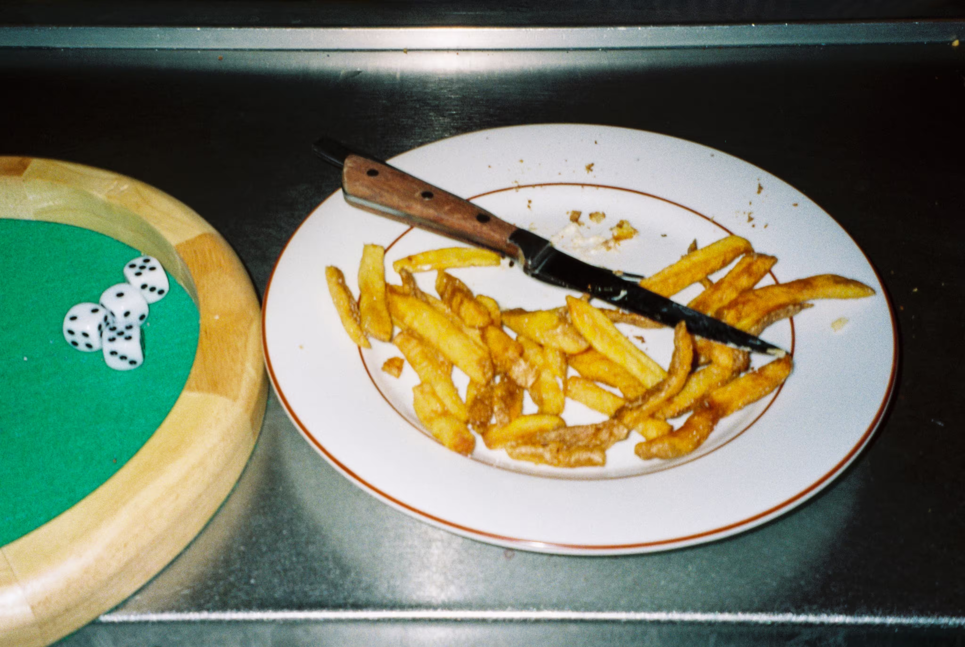 Plate with leftover French fries and a knife on a metal surface next to a wooden dice tray holding five dice.