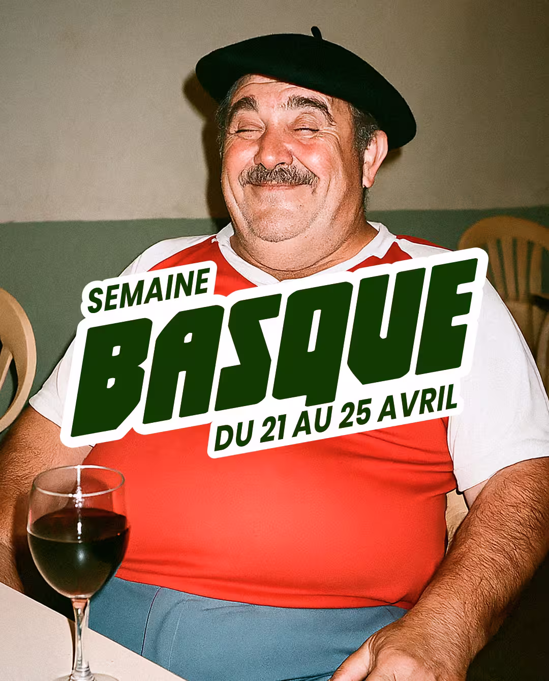 Homme souriant portant un béret noir et un maillot rouge et blanc, assis avec un verre de vin rouge devant lui.