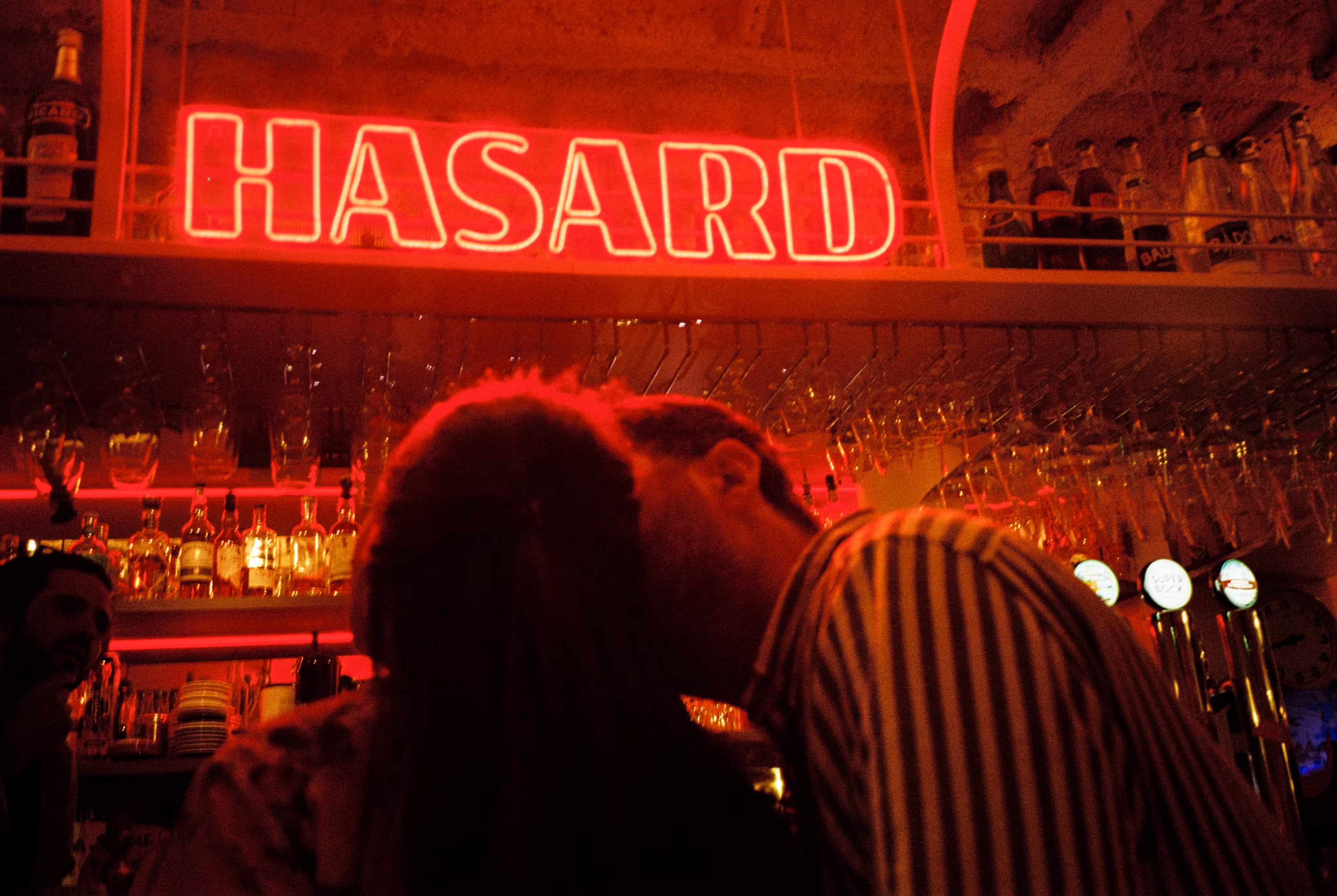 Un couple s'embrasse dans un bar éclairé par une enseigne néon rouge affichant le mot « HASARD ».